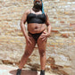 Person wearing a black bra and gaff set with a blue headscarf against a brick wall.